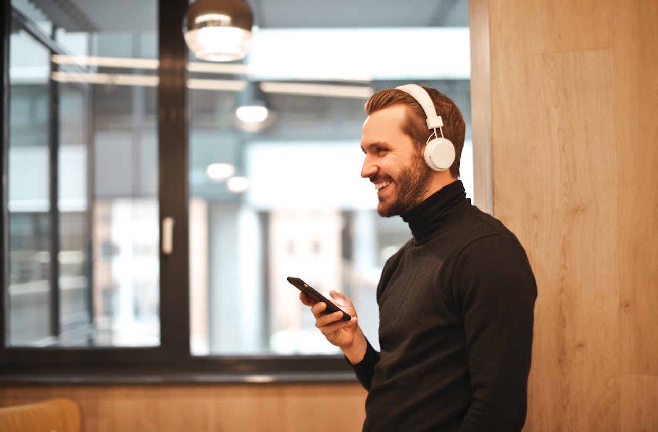 A cheerful man in a black turtleneck enjoys music on headphones, holding a smartphone indoors.